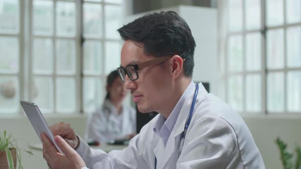 Asian Man Doctor Is Using Tablet While Work With Desktop Computer In Workplace. Medical Concept