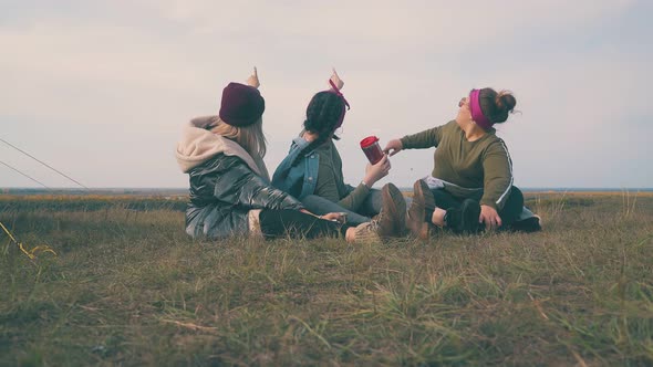 Happy Women with Drinks Look at Sky Sitting on Grass By Tent