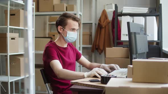 Man Employee of Warehouse Wearing Medical Mask and Protective Gloves Holding Delivery Box and Typing