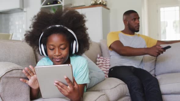 African american daughter wearing headphones using tablet while her father watching tv