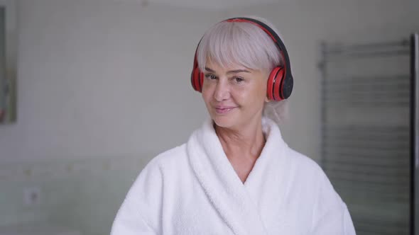 Portrait of Greyhaired Middle Aged Woman in White Bathrobe and Headphones Posing in Bathroom at Home