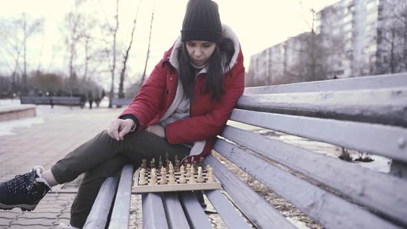 Young Woman in Winter Clothes Plays Chess Sitting on Bench in City Park