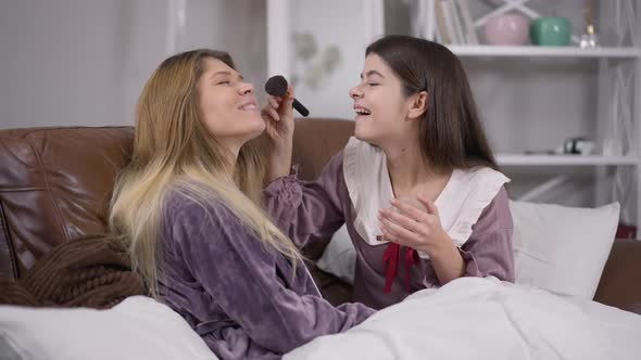 Joyful Pretty Woman Laughing Applying Face Powder Resting with Friend at Pajama Party Indoors