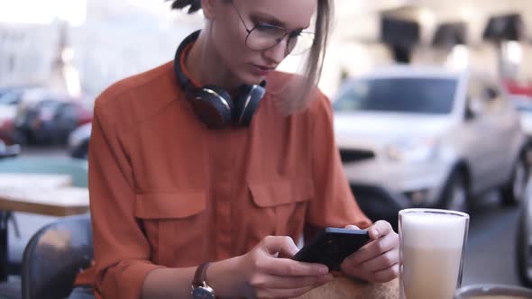 An Attractive Blonde Girl with Hair in a Bundle and Glasses is Sitting in a Cafe on the Street Next