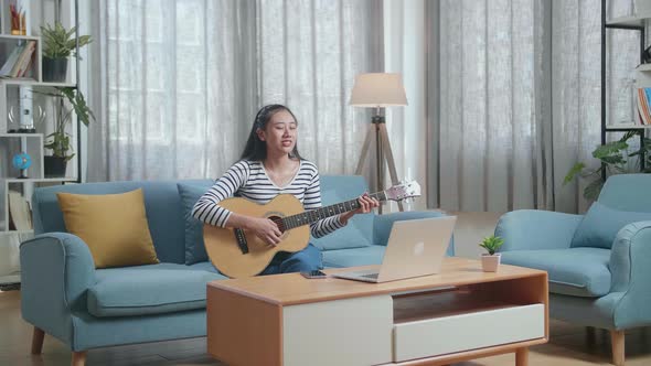 Asian Woman With Laptop Singing And Playing Guitar At Home