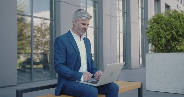 Mature Business Man Sitting on Bench and Working with Laptop in City Street