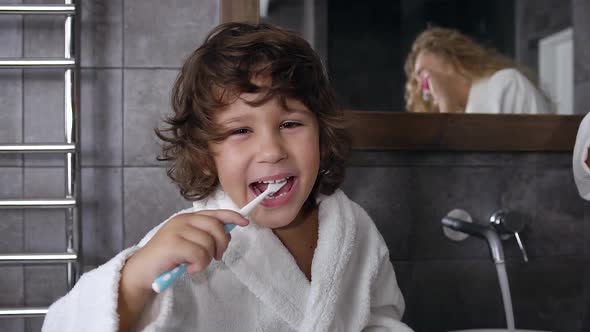 Little Boy with Dark Curly Hair Dressed in Robe Brushing His Teeth