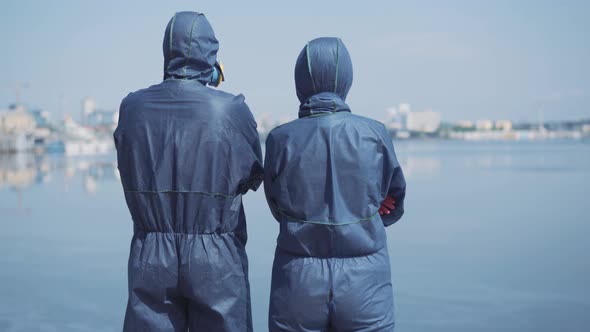 Back View of Two People in Biohazard Suits Standing at Riverbank and Looking at Blurred City on the