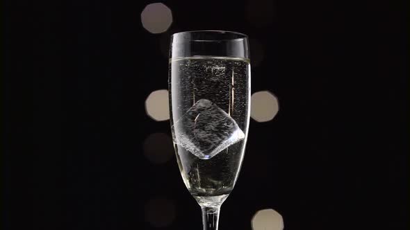 Cube of Ice Swirls in a Glass of Champagne. Bokeh Blinking Black Background. Close Up