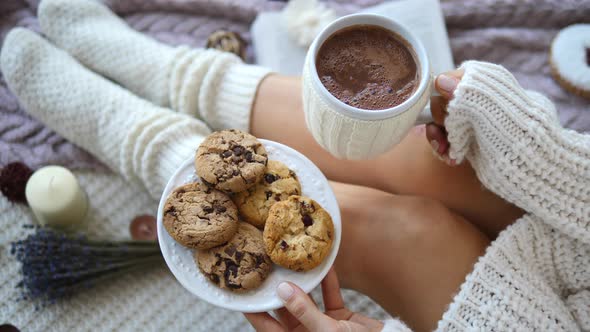 Trendy Girl In Knitted Sweater And Socks Holding Hot Chocolate And Cookies At Home