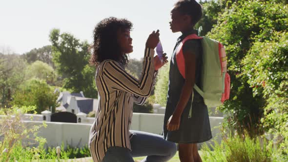 African american mother putting on face mask on daughter with backpack outdoors on a bright sunny da