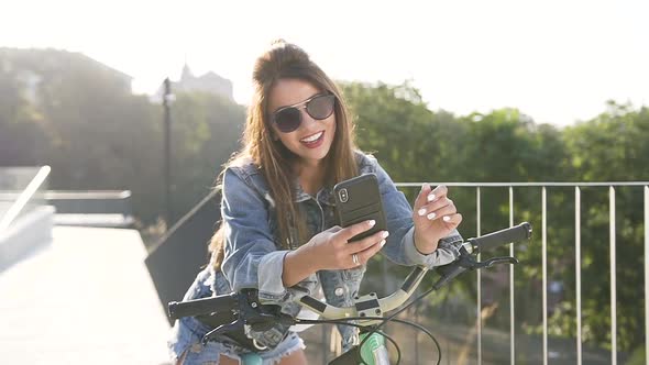 Young Woman in Sunglasses, Dressed in Jeans Clothing Walking with a Bicycle at the Park