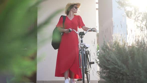 Wide Shot Confident Young Woman in Red Dress Sunglasses and Straw Hat with Bike Standing on Porch