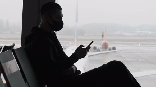 Masked Young Man in Airport Waiting Room Sits and Using a Smartphone By Window