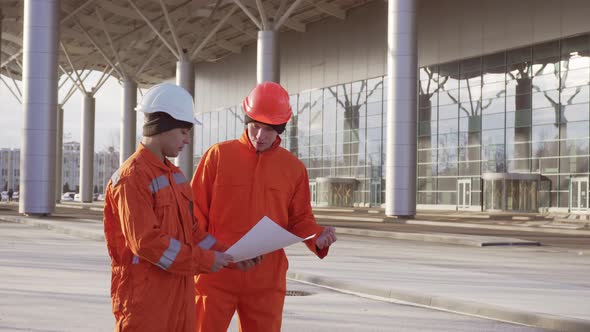 Two Construction Workers in Orange Uniform and Hardhats Examining the Constructed Building Together