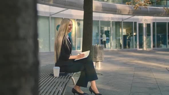 Successful Young Woman Taking Notes Outdoors. Side View of Elegant Businesswoman Sitting on Bench in