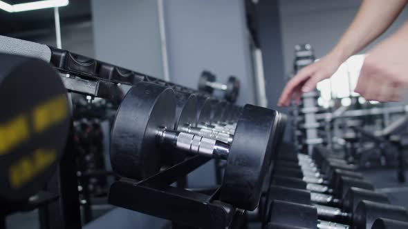 Male athlete with fitness bracelet on wrist approaches rack with dumbbells and takes one in hand