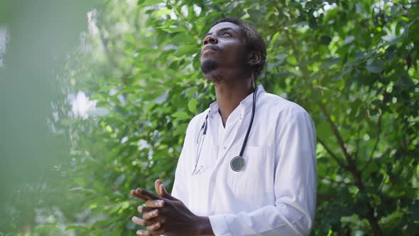 Young African American Man in Doctor Uniform Standing Outdoors on Sunny Day Praying in Slow Motion