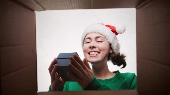 Portrait of a young happy woman in a Christmas hat opening a parcel with a gift.