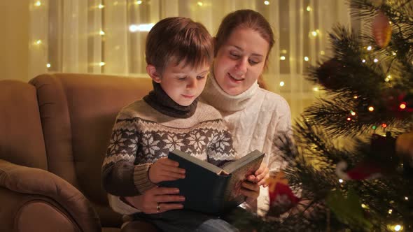 Portrait of Smiling Mother with Son Reading Christmas Story Book Next to Glowing Christmas Tree