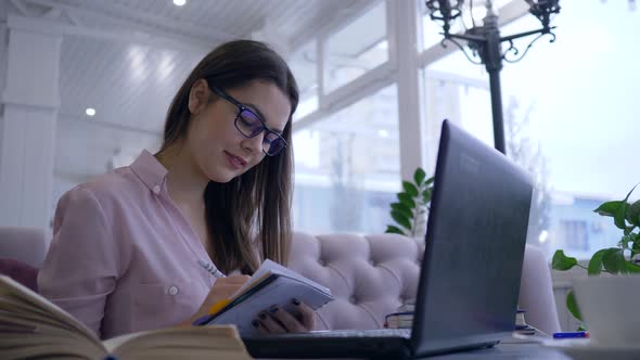 Education in Internet, Happy Girl Student in Eyeglasses Writes Notes in Notebook Sitting at Table