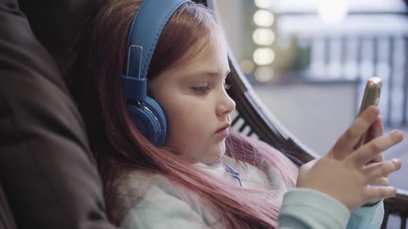 Side View Close-up of Sad Little Girl in Headphones Using Smartphone Indoors. Portrait of Upset