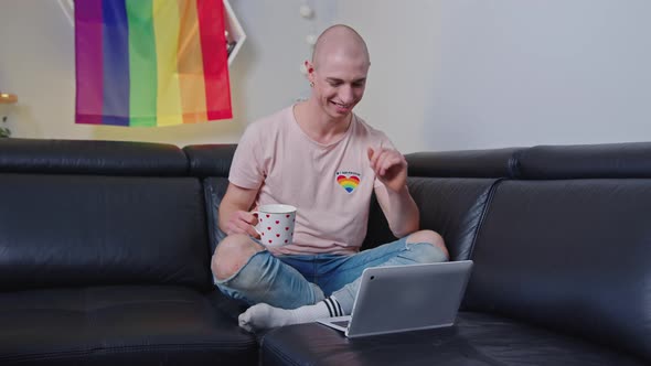 Happy Smiling Homosexual Bald Caucasian Man Sitting on a Black Couch Holding a Cup of Tea or Coffee
