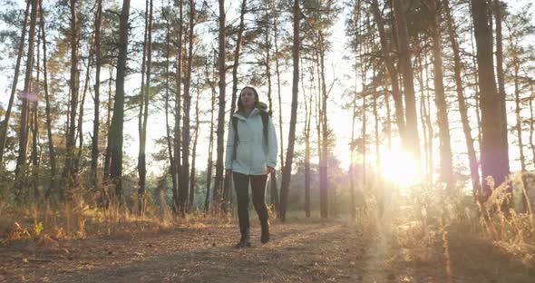 Happy female traveler in jacket with tourist backpack walking in autumn forest at sunset sunlight