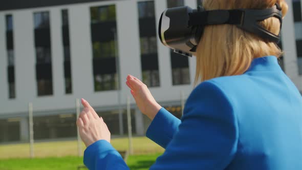 A Happy Woman in the Suit is Standing Outside and Gestures in Augmented Reality in 3D VR Goggles