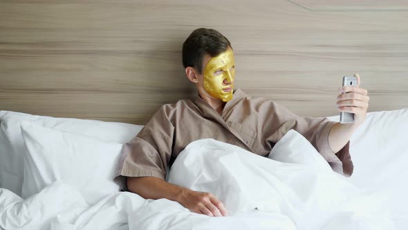 Guy with Golden Face Mask Makes Selfie in Soft Bed in Hotel