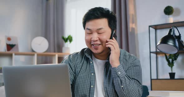 Chinese Man which Sitting in front of Computer at Home Office and Talking on Mobile