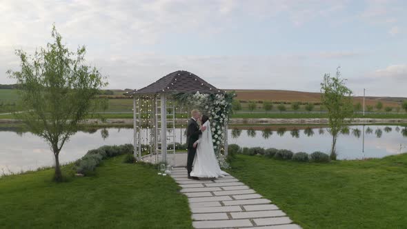 Newlyweds Groom Bride Embracing Huggings Kissing Wedding Matrimony Ceremony Arch with Flowers