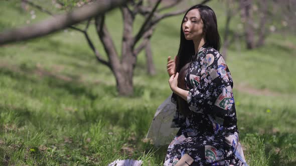 Side View of Confident Beautiful Asian Young Woman in Kimono Sitting on Green Spring Meadow in Park