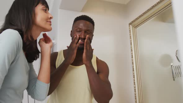 Happy diverse couple looking in mirror and using cream in bathroom