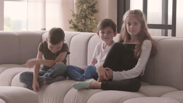 Portrait of Cute Older Sister and Two Younger Adorable Brothers Sitting on the Couch Looking