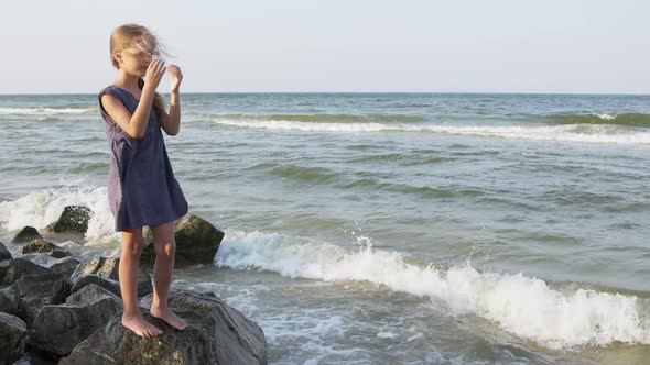 A Girl in a Blue Dress with Polka Dots and Sunglasses Looks Into the Distance on the Beach