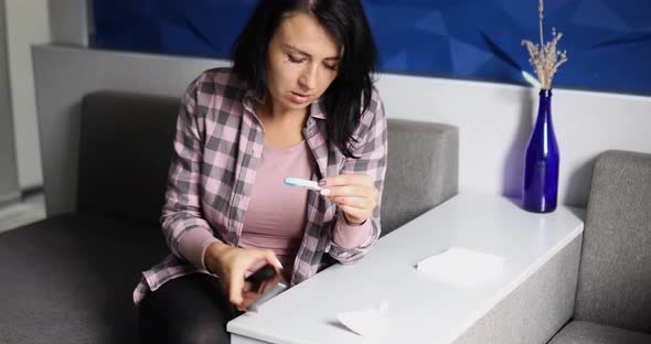 Ill lady wiping nose and looking at electronic thermometer, take phone to call doctor