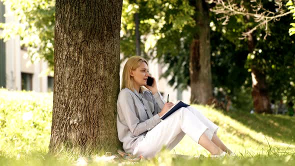 Student Girl Sitting on a Grass Near a Tree and Talking on the Phone