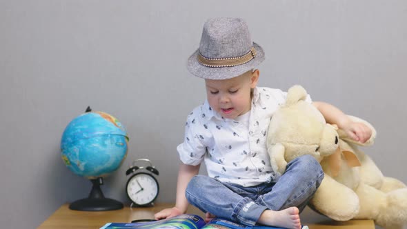 Happy Baby Boy in Hat and Jeans Holding Teddy Bear and Looking Into Book Smile