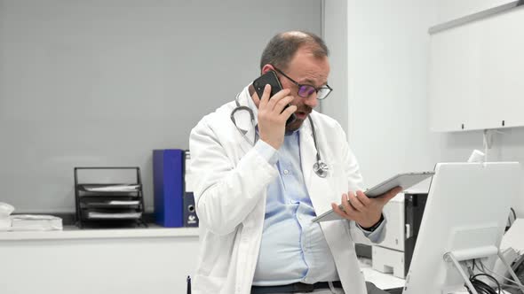 Male Physician with Stethoscope Sitting at Desk Talking on Mobile Phone and Looking at Digital