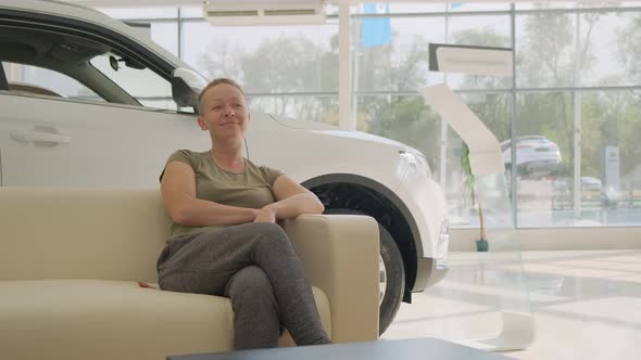 Happy woman in showroom in anticipation of deal sits on couch in front of new car