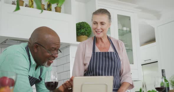 Smiling senior diverse couple wearing blue aprons and using tablet in kitchen