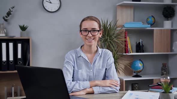 Woman in Glasses which Posing on Camera with Sincerely Smile at Her Workplace with Computer