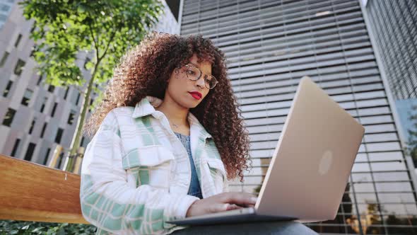 Young Female Freelancer with Afro Hairstyle is Sitting Outdoors on Street in the City in Business