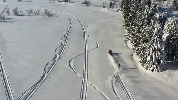 Aerial view of snowmobile scooter towing man on skis by conifer in snowy winter mountain landscape,