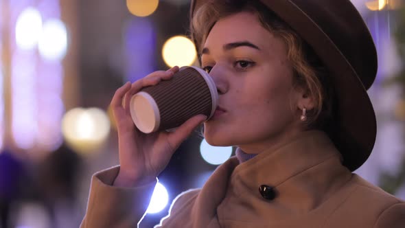 Beautiful Woman in Hat and Coat Drinks Coffee in the Night City