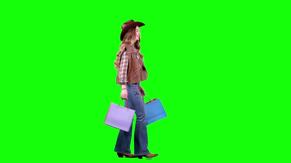 Girl in a Hat and Cowboy Boots Comes with Bags in Her Hands. Green Screen. Side View