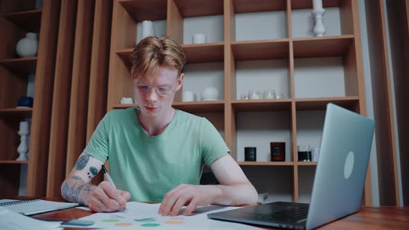 Guy Studying at Home Looks at a Laptop Writes in Notebook and Reads a Textbook