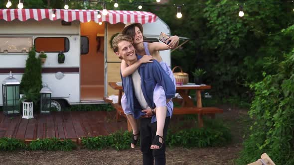 Happy Young Lovely Couple Having Fun Near Trailer in the Park Man Piggybacking Girl