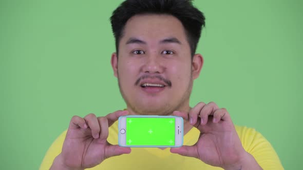 Face of Happy Young Overweight Asian Man Thinking While Showing Phone